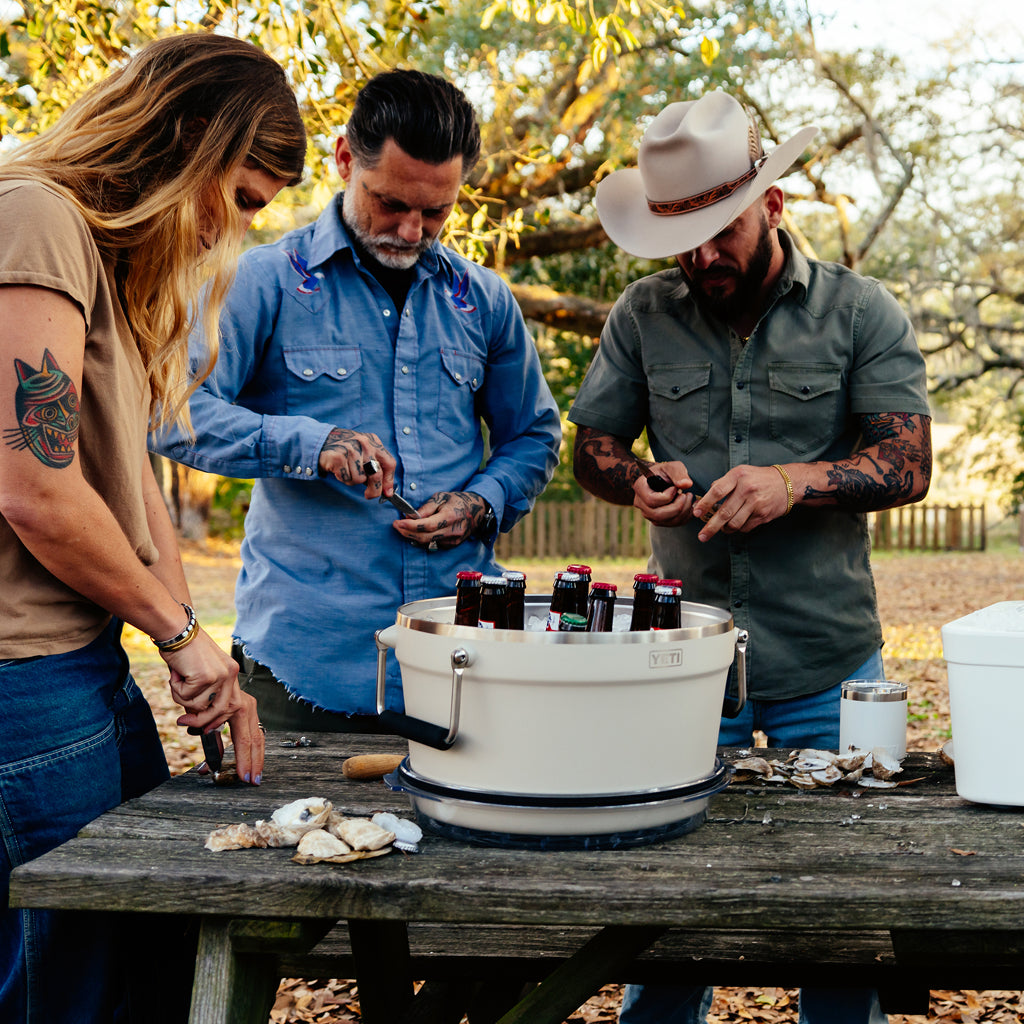 Three people preparing food outdoors with a beverage tub full of drinks on a wooden table.