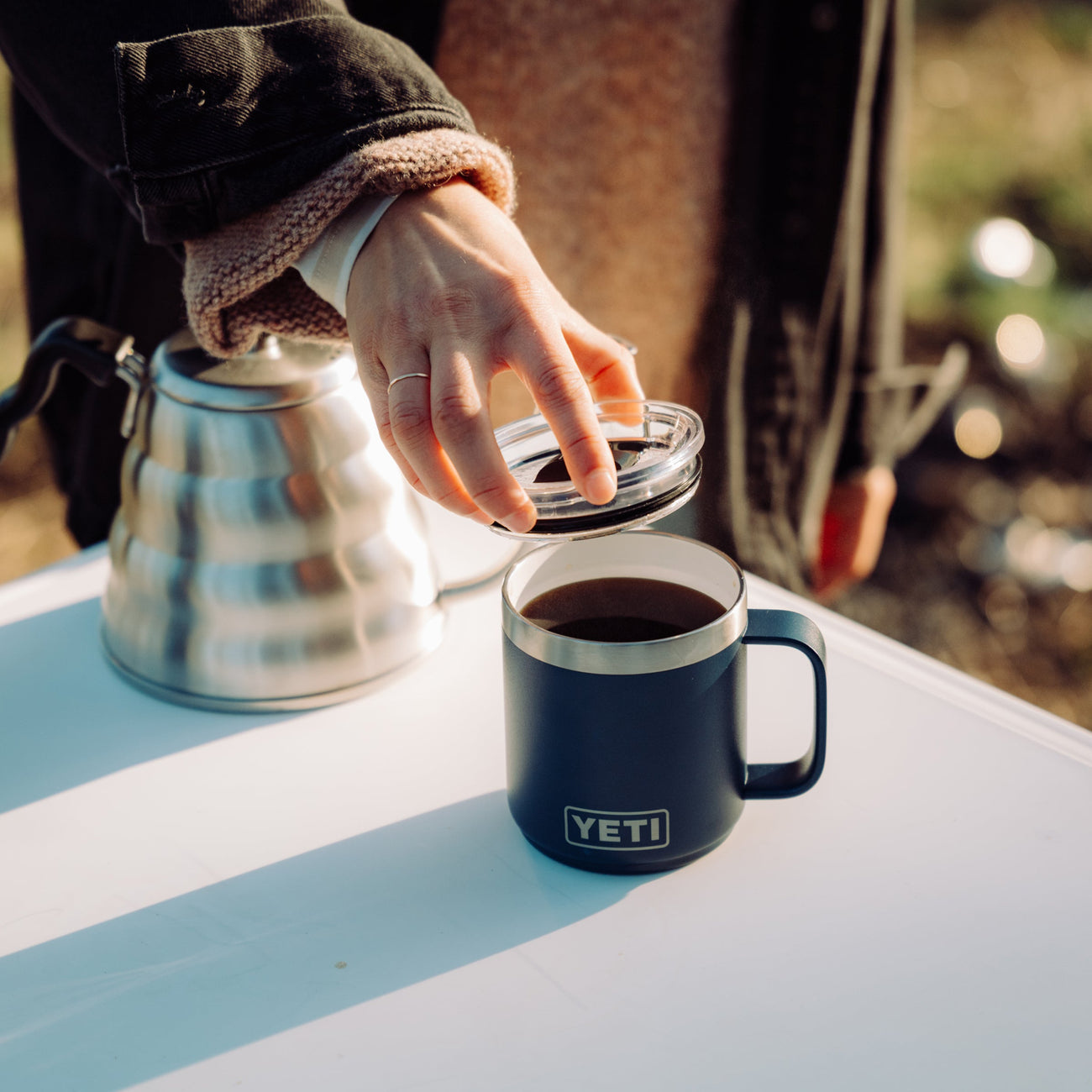 Hand opening a YETI mug filled with coffee on a table outdoors.