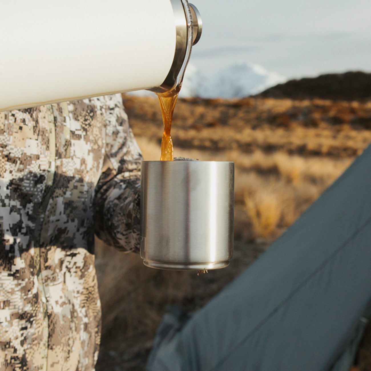 Person pouring a hot beverage into a metal cup with a scenic background