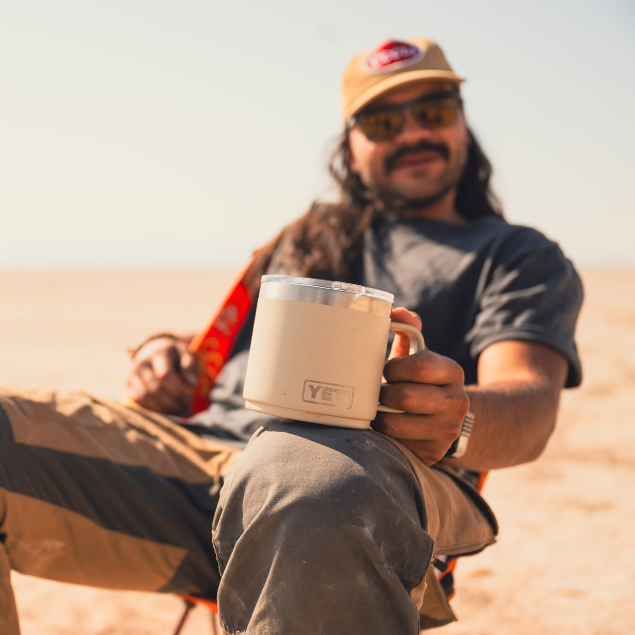 Man sitting outdoors holding a cape taupe mug with 'YETI' branding, wearing a cap and sunglasses.
