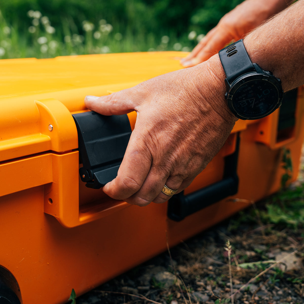 A close-up of a person's hands opening the latch of a bright orange hard-sided case outdoors, with grass and dirt visible in the background.
